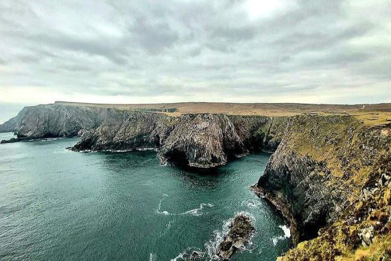 Een ruige kustlijn met steile kliffen kijkt uit over een kalme zee onder een bewolkte lucht. De kliffen, bedekt met stukken gras, steken dramatisch uit in het water en creëren natuurlijke formaties die perfect zijn voor een rondreis Ierland. De oceaan is diepblauwgroen en golven klotsen zachtjes tegen de rotsachtige kust. | Echt Ierland Vakanties