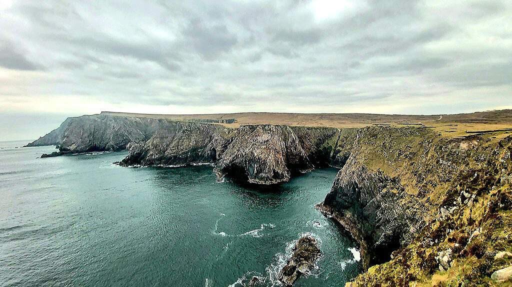 Een ruige kustlijn met steile kliffen kijkt uit over een kalme zee onder een bewolkte lucht. De kliffen, bedekt met stukken gras, steken dramatisch uit in het water en creëren natuurlijke formaties die perfect zijn voor een rondreis Ierland. De oceaan is diepblauwgroen en golven klotsen zachtjes tegen de rotsachtige kust. | Echt Ierland Vakanties