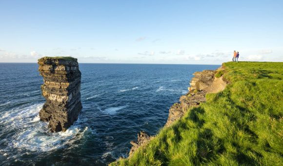 Een hoge zeestapel rijst op uit de oceaan bij een met gras begroeide klifrand. Twee mensen staan op de klif, ondergedompeld in hun rundreise Irland, en staren naar de stapel. De helderblauwe lucht en kalme zee creëren een vredig kustlandschap terwijl golven zachtjes tegen de basis van de rotsformatie beuken. | Echt Ierland Vakanties