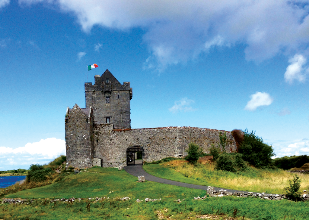 Een stenen kasteel met een hoge toren en kantelen staat op een met gras begroeide heuvel onder een blauwe lucht en nodigt reizigers uit voor hun rondreis Ierland. Een Ierse vlag wappert boven op de toren. Het bouwwerk is omringd door een korte stenen muur en een gewelfde ingang leidt naar een pad door het groen terwijl er dunne wolken in de lucht drijven. | Echt Ierland Vakanties