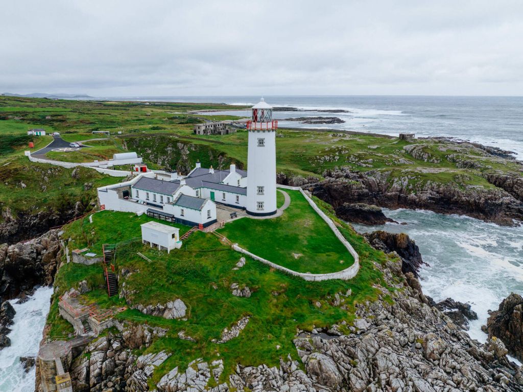Een witte vuurtoren met een rode top staat op een rotsachtige groene klif, en biedt het perfecte uitkijkpunt voor degenen die een rondreis Ierland maken. Omringende gebouwen en een stenen pad leiden naar de waterkant. De kustlijn strekt zich uit tot in de verte onder een bewolkte lucht terwijl golven tegen de rotsen eronder beuken. | Echt Ierland Vakanties