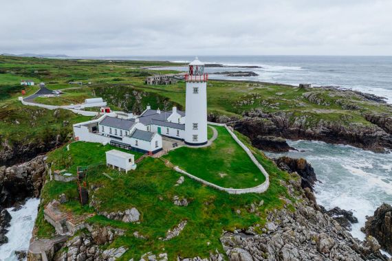 Een witte vuurtoren met een rode top staat op een rotsachtige groene klif, en biedt het perfecte uitkijkpunt voor degenen die een rondreis Ierland maken. Omringende gebouwen en een stenen pad leiden naar de waterkant. De kustlijn strekt zich uit tot in de verte onder een bewolkte lucht terwijl golven tegen de rotsen eronder beuken. | Echt Ierland Vakanties