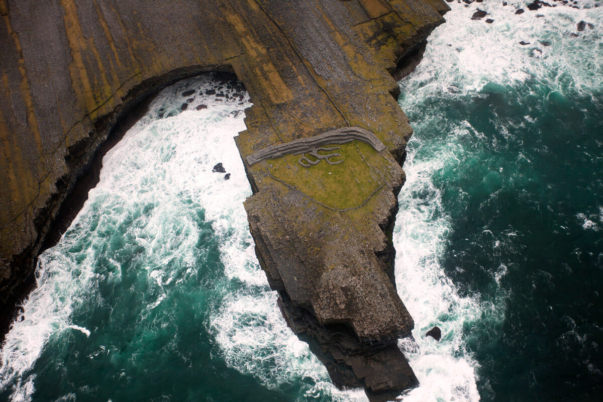 Luchtfoto van ruige kliftoppen die het woeste oceaanwater ontmoeten: een perfect tafereel voor degenen die een urlaub Ierland overwegen. De kliffen, met gelaagde rotsformaties en groene mosplekken, zijn getuige van witte golven die krachtig op hun basis beuken, wat het opvallende contrast tussen aarde en water laat zien. | Echt Ierland Vakanties