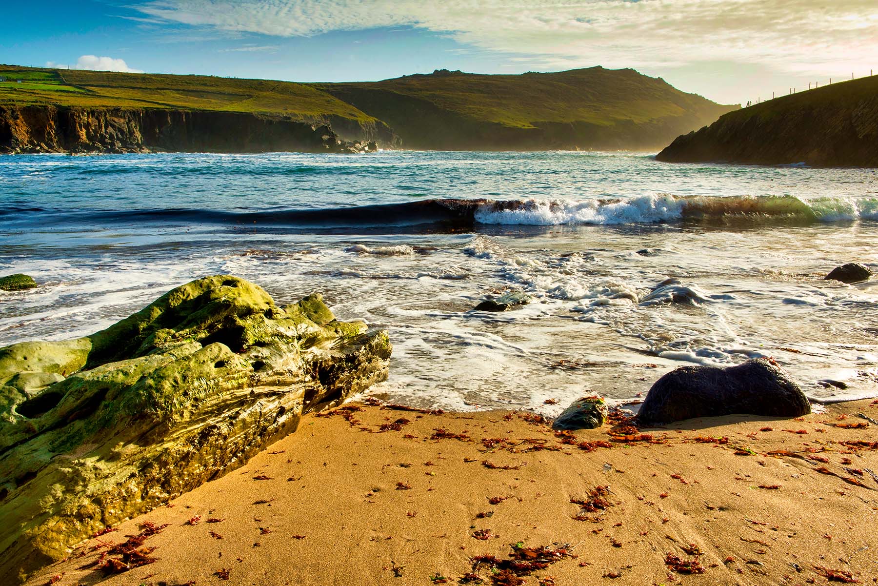Ontdek de schilderachtige kustlijn van Ierland, waar een goudkleurig zandstrand bezaaid is met zeewier en omspoeld door zachte golven. Een met mos bedekte rots steekt af tegen ruige kliffen en met gras begroeide heuvels. Ervaar deze schoonheid tijdens uw rondreis Ierland onder een gedeeltelijk bewolkte hemel, met zonlicht dat het landschap verlicht. | Echt Ierland Vakanties
