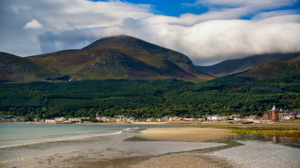 Een schilderachtig kustlandschap in Ierland met een zandstrand met zachte golven aan de linkerkant. Op de achtergrond omlijsten weelderige groene heuvels en bergen, deels bedekt door laaghangende wolken, een klein stadje. Ervaar dit charmante uitzicht op uw rondreis Ierland onder een helderblauwe lucht. | Echt Ierland Vakanties
