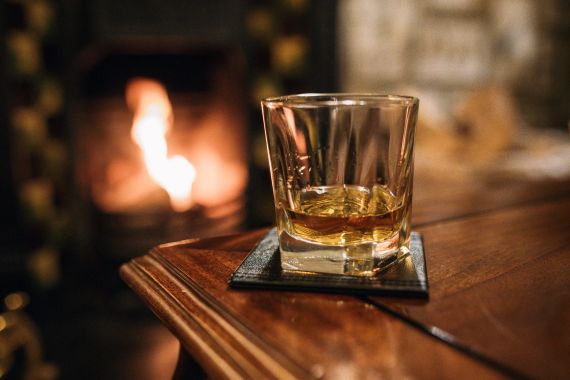 Een glas whisky staat op een onderzetter op een houten tafel, met de gezellige ambiance van een Ierse pub. De kamer is warm verlicht, wat doet denken aan een onvergetelijke reis in Ierland. Vlammen in de open haard werpen een flikkerende gloed, die subtiel weerkaatst op het oppervlak van het glas. | Echt Ierland Vakanties