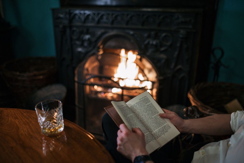 Een persoon zit voor een brandende open haard en leest een boek. De afbeelding legt de gezellige ambiance vast die doet denken aan een vakantie in Ierland, met een houten tafel op de voorgrond met een glas whisky. Twee rieten manden staan naast de open haard en verrijken deze warme, uitnodigende scène. | Echt Ierland Vakanties