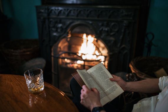 Een persoon zit voor een brandende open haard en leest een boek. De afbeelding legt de gezellige ambiance vast die doet denken aan een vakantie in Ierland, met een houten tafel op de voorgrond met een glas whisky. Twee rieten manden staan naast de open haard en verrijken deze warme, uitnodigende scène. | Echt Ierland Vakanties