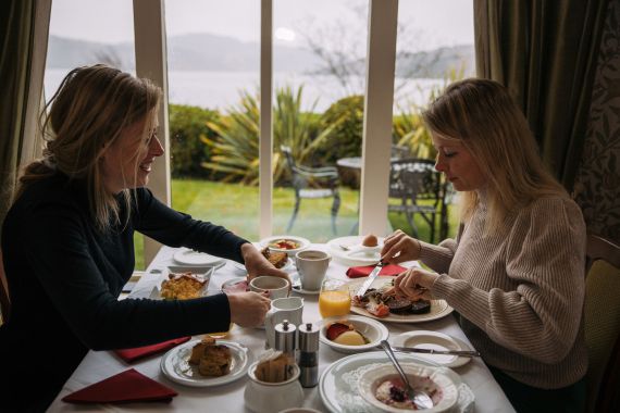 Twee mensen genieten van hun ontbijt aan een tafel bij een groot raam met uitzicht op een tuin en meer tijdens hun urlaub Irland. De tafel heeft borden met verschillende soorten eten, waaronder fruit, gebak en eieren. De setting oogt gezellig, met natuurlijk licht dat binnenstroomt terwijl ze van hun maaltijden genieten. | Echt Ierland Vakanties