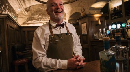 Een lachende barman met een wit overhemd, bruine vlinderdas en groene schort staat achter een houten bar in een rustieke Ierse pub. Zijn witte baard contrasteert met de schemerige, gewelfde plafonds. Flessen staan op de voorgrond in warme verlichting: een perfect tafereel voor uw urlaub Irland-avontuur. | Echt Ierland Vakanties
