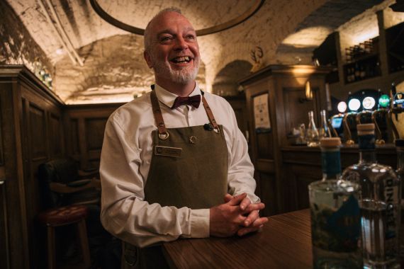 Een lachende barman met een wit overhemd, bruine vlinderdas en groene schort staat achter een houten bar in een rustieke Ierse pub. Zijn witte baard contrasteert met de schemerige, gewelfde plafonds. Flessen staan op de voorgrond in warme verlichting: een perfect tafereel voor uw urlaub Irland-avontuur. | Echt Ierland Vakanties