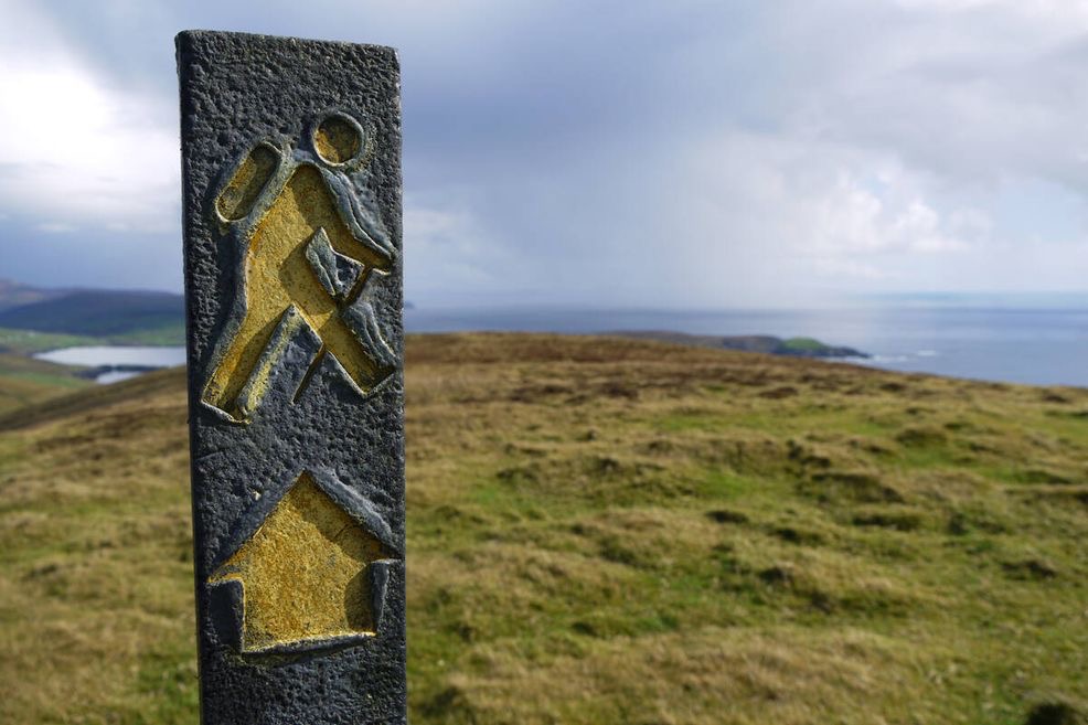 Een verweerde routemarkering met een wandelaarsymbool en pijl staat op de voorgrond en wijst de weg. Op de achtergrond strekken glooiende groene heuvels zich uit naar een rustige zee onder een bewolkte lucht, wat doet denken aan een kustwandelpad dat perfect is voor mensen die op vakantie zijn in Ierland. | Echt Ierland Vakanties