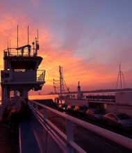 Een veerbootdek tijdens zonsondergang in Ierland met een levendige lucht van oranje en roze tinten. Passagiers, waaronder een die het moment vastlegt voor hun reisen irland herinneringen, zijn zichtbaar tussen geparkeerde auto's. Het silhouet van de structuur van de veerboot steekt af tegen de kleurrijke lucht. | Echt Ierland Vakanties