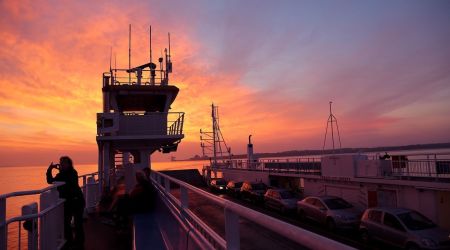 Een veerbootdek tijdens zonsondergang in Ierland met een levendige lucht van oranje en roze tinten. Passagiers, waaronder een die het moment vastlegt voor hun reisen irland herinneringen, zijn zichtbaar tussen geparkeerde auto's. Het silhouet van de structuur van de veerboot steekt af tegen de kleurrijke lucht. | Echt Ierland Vakanties
