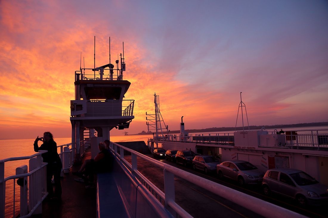 Een veerbootdek tijdens zonsondergang in Ierland met een levendige lucht van oranje en roze tinten. Passagiers, waaronder een die het moment vastlegt voor hun reisen irland herinneringen, zijn zichtbaar tussen geparkeerde auto's. Het silhouet van de structuur van de veerboot steekt af tegen de kleurrijke lucht. | Echt Ierland Vakanties