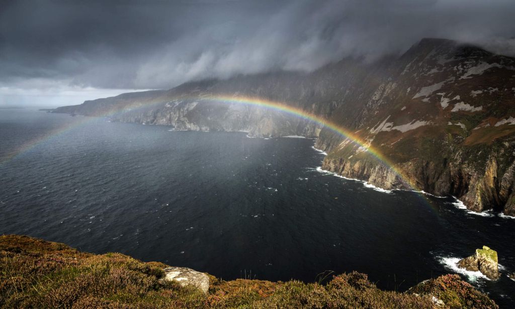 Een dramatisch kustlandschap in Ierland toont steile kliffen die een donkere, woelige zee ontmoeten onder een stormachtige lucht. Een levendige regenboog buigt van de zee naar de kliffen en voegt kleur toe aan het sfeervolle tafereel. Dit wilde terrein, ideaal voor een rondreis Ierland, heeft stukken groene vegetatie onder zware wolken. | Echt Ierland Vakanties