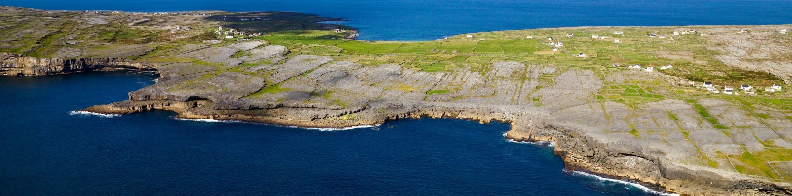 Luchtfoto van een rotsachtige kustlijn in Ierland vakanties, met stukken groene velden onder een helderblauwe lucht. De kustlijn buigt zachtjes af naar de diepblauwe zee, ver land aan de overkant van het water en verspreide kleine gebouwen die het landschap bezaaien. Perfect voor een rundreise Ierland. | Echt Ierland Vakanties