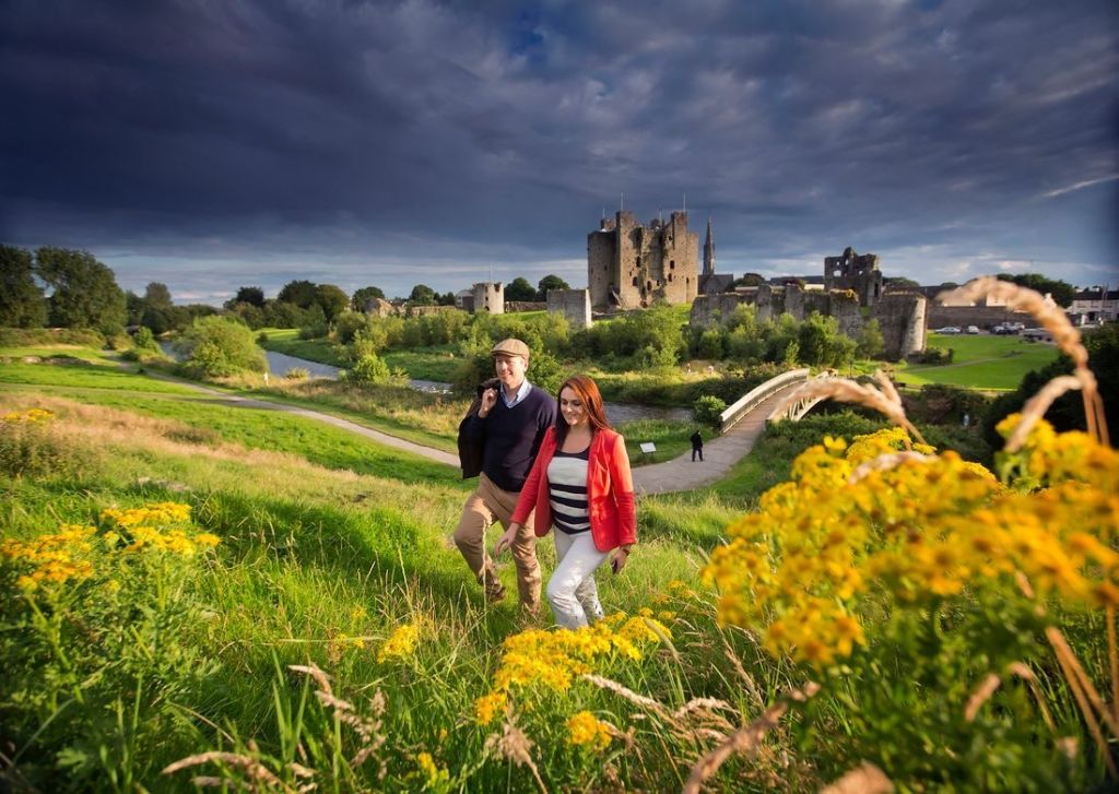 Een man en een vrouw wandelen door een weelderig groen veld met levendige gele bloemen op de voorgrond. Achter hen staat een historisch kasteel tegen een dramatische bewolkte lucht. Vlakbij hen steekt een stenen brug een rivier over. Ze omarmen de charme van "vakantie Ierland" en lijken gelukkig en ontspannen in casual kleding. | Echt Ierland Vakanties