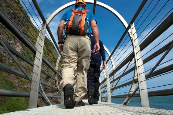 Twee mensen lopen over een metalen hangbrug met gebogen steunen. De persoon op de voorgrond draagt een helm, oranje rugzak en beige broek. Links is een rotsachtige heuvel te zien en rechts is de zee zichtbaar onder een helderblauwe lucht. Perfect voor iedereen die van een vakantie in Ierland houdt. | Echt Ierland Vakanties