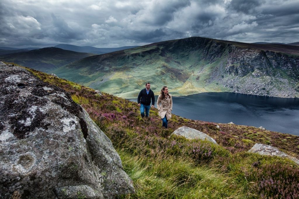 Een man en een vrouw wandelen een met gras begroeide heuvel op, met grote rotsblokken op de voorgrond. Deze beelden symboliseren de geest van 'Urlaub in Ierland'. Het donkere, rustige meer tegen de achtergrond van ruige groene heuvels en een dramatische lucht duiden op een naderende storm, wat een gevoel van avontuur en natuurlijke schoonheid oproept. | Echt Ierland Vakanties