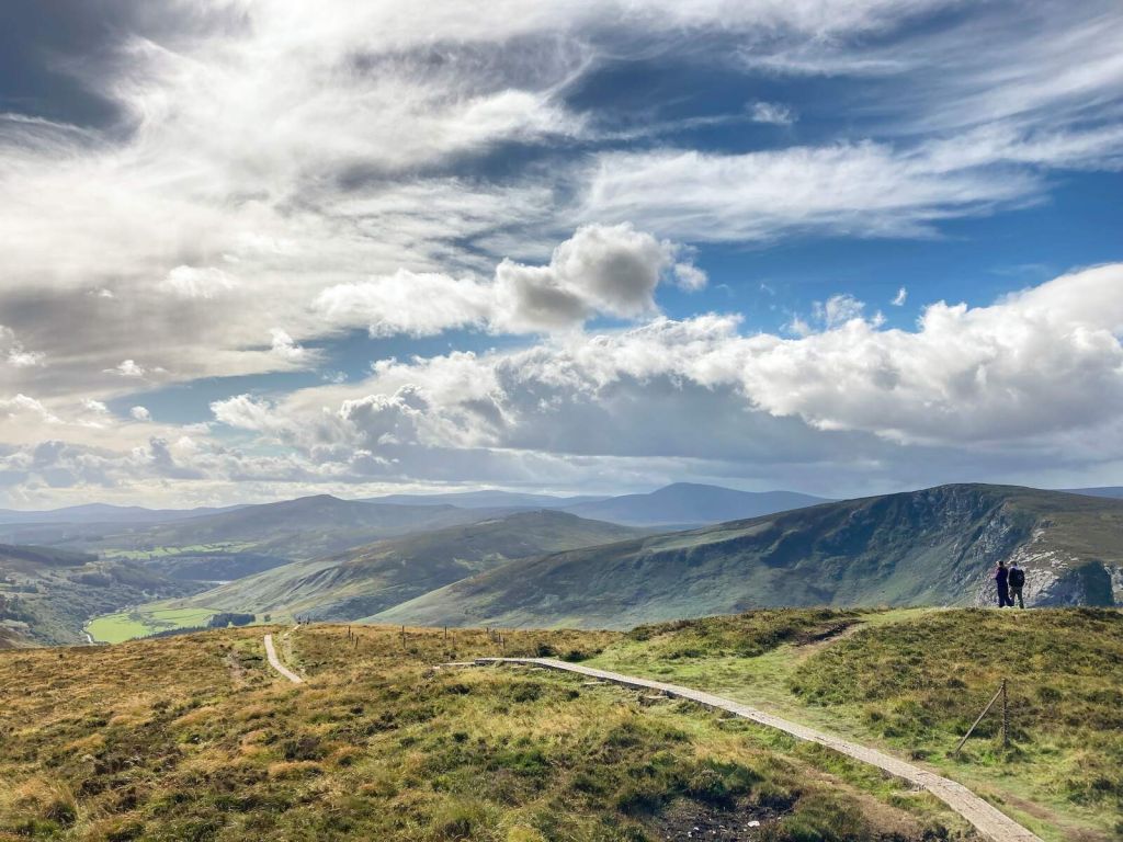 Een schilderachtig uitzicht op een uitgestrekt heuvellandschap onder een gedeeltelijk bewolkte lucht in het hart van een rundreise Ierland. Twee mensen staan op een pad, omringd door weelderig groen gras en verre bergen. Een kronkelend pad leidt naar de horizon terwijl zonlicht door de wolken breekt en licht en schaduw op het terrein werpt. | Echt Ierland Vakanties