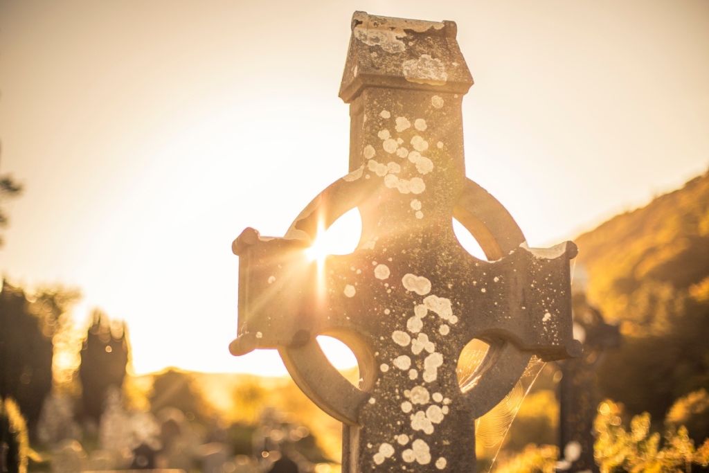 Een verweerd stenen Keltisch kruis staat op een begraafplaats, afgetekend tegen de gouden gloed van een ondergaande zon. Dit pittoreske tafereel wordt vaak ervaren tijdens een rondreis Ierland, waar wazige bomen en heuvels baden in warm licht, wat een serene en nostalgische sfeer creëert. | Echt Ierland Vakanties