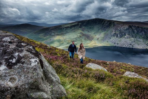 Een man en een vrouw wandelen een met gras begroeide heuvel op, met grote rotsblokken op de voorgrond. Deze beelden symboliseren de geest van 'Urlaub in Ierland'. Het donkere, rustige meer tegen de achtergrond van ruige groene heuvels en een dramatische lucht duiden op een naderende storm, wat een gevoel van avontuur en natuurlijke schoonheid oproept. | Echt Ierland Vakanties