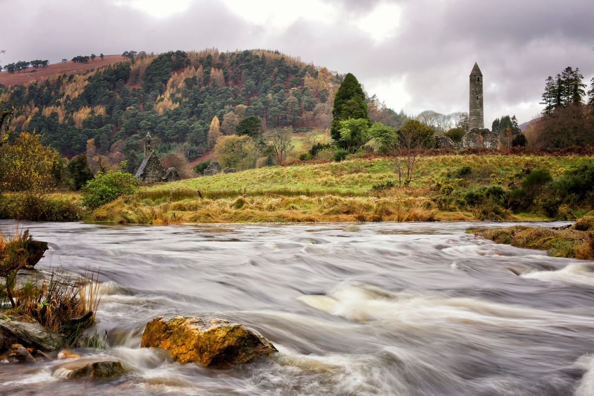 Een rivier met snelstromend water kruist de voorgrond, omzoomd door grasachtige oevers. Op de achtergrond staat een hoge, ronde middeleeuwse toren omringd door bomen, met een heuvelachtig landschap en een bewolkte lucht die het tafereel compleet maken. Vroege herfstkleuren stippelen de bomen - een perfect momentopname van vakantie Ierland. | Echt Ierland Vakanties