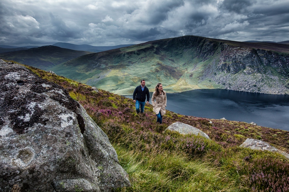 Een man en een vrouw wandelen een met gras begroeide heuvel op, met grote rotsblokken op de voorgrond. Deze beelden symboliseren de geest van 'Urlaub in Ierland'. Het donkere, rustige meer tegen de achtergrond van ruige groene heuvels en een dramatische lucht duiden op een naderende storm, wat een gevoel van avontuur en natuurlijke schoonheid oproept. | Echt Ierland Vakanties