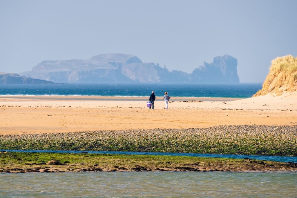 Twee mensen slenteren langs een zandstrand met verre rotsachtige eilanden als achtergrond op hun rundreise Irland. De voorgrond toont groene met zeewier bedekte gebieden en ondiep water, terwijl de helderblauwe lucht de rust versterkt. Met vegetatie bedekte duinen zijn rechts van de scène zichtbaar. | Echt Ierland Vakanties