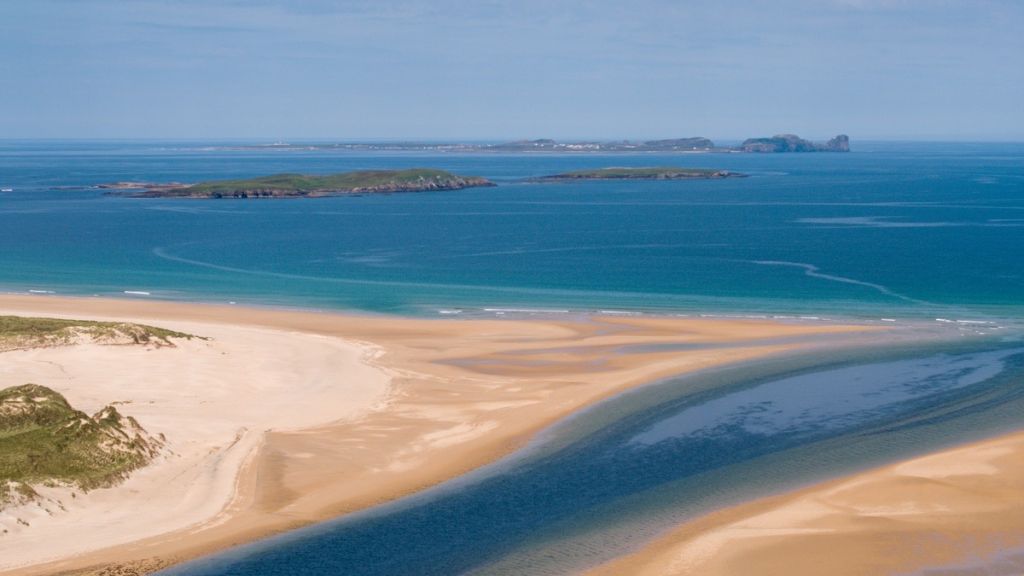 Een luchtfoto van een adembenemend kustlandschap dat doet denken aan een vakantie in Ierland, met een zandstrand met duinformaties. Een smalle, donkerblauwe rivier slingert door het zand om de serene turquoise oceaan te ontmoeten, terwijl kleine groene eilandjes de horizon bedekken onder een helderblauwe lucht. | Echt Ierland Vakanties