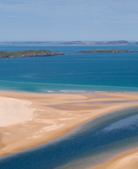 Een luchtfoto van een adembenemend kustlandschap dat doet denken aan een vakantie in Ierland, met een zandstrand met duinformaties. Een smalle, donkerblauwe rivier slingert door het zand om de serene turquoise oceaan te ontmoeten, terwijl kleine groene eilandjes de horizon bedekken onder een helderblauwe lucht. | Echt Ierland Vakanties