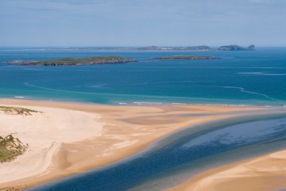 Een luchtfoto van een adembenemend kustlandschap dat doet denken aan een vakantie in Ierland, met een zandstrand met duinformaties. Een smalle, donkerblauwe rivier slingert door het zand om de serene turquoise oceaan te ontmoeten, terwijl kleine groene eilandjes de horizon bedekken onder een helderblauwe lucht. | Echt Ierland Vakanties