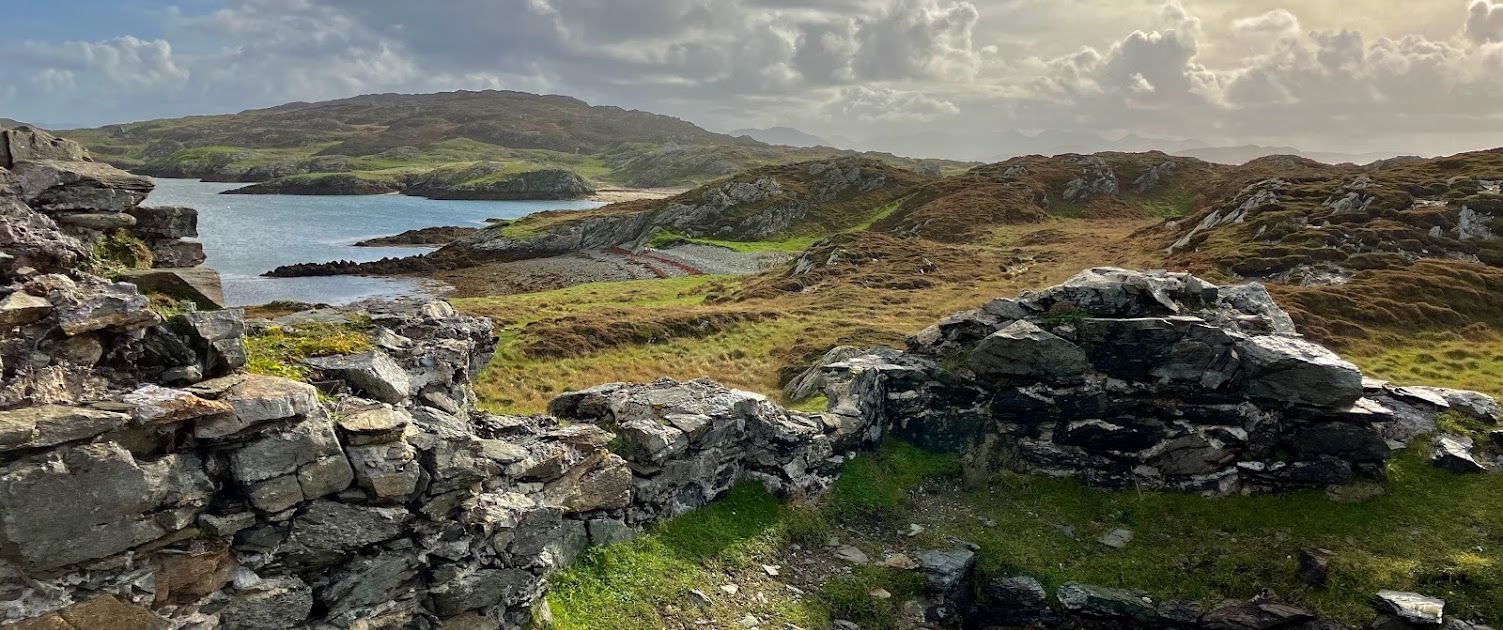 Uitzicht vanaf een stenen ruïne met uitzicht op een schilderachtig landschap met een kalm water, perfect voor degenen die een rondreis Ierland maken. Met mos bedekte stenen stippelen de voorgrond, terwijl met gras begroeide heuvels zich uitstrekken tot de achtergrond. De gedeeltelijk bewolkte lucht laat zacht zonlicht zachte schaduwen werpen over dit betoverende terrein. | Echt Ierland Vakanties