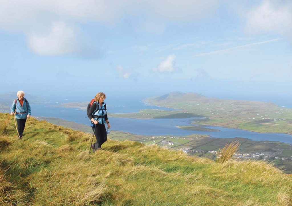 Twee mensen wandelen op een grashelling met rugzakken, uitkijkend over een uitgestrekt landschap. De achtergrond toont een uitgestrekt uitzicht op weelderige groene velden, wat doet denken aan een onvergetelijke vakantie in Ierland, met een groot waterlichaam en verre heuvels onder een gedeeltelijk bewolkte lucht. De scène straalt avontuur en natuurlijke schoonheid uit. | Echt Ierland Vakanties