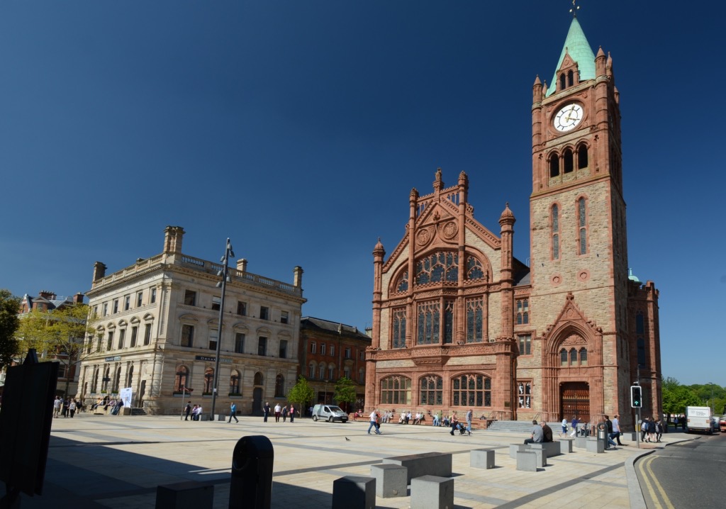 Een groot plein in het hart van uw rundreise Ierland toont mensen die lopen en zitten. Rechts staat een groot stenen gebouw met een klokkentoren en boogramen, aan de linkerkant aangevuld met een kleiner klassiek zuilengebouw. De helderblauwe lucht werpt fel zonlicht over dit charmante tafereel. | Echt Ierland Vakanties