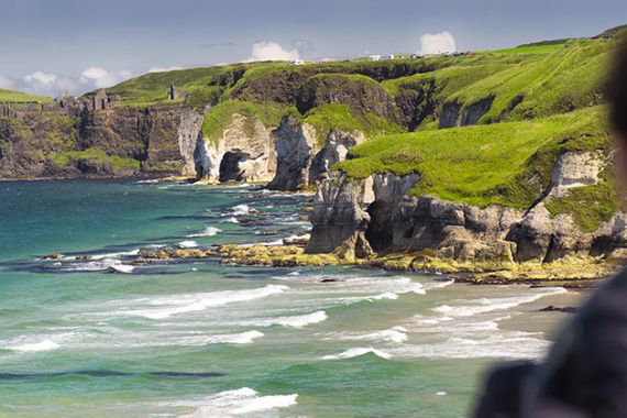 Een ruige kustlijn met steile, met gras begroeide kliffen en rotsformaties strekt zich uit langs een levendige blauwe zee, perfect voor een onvergetelijke vakantie in Ierland. Zachte golven raken de kust bij oude ruïnes bovenop de kliffen. Een wazig figuur op de voorgrond staart naar dit serene tafereel onder een heldere lucht bezaaid met wolken. | Echt Ierland Vakanties