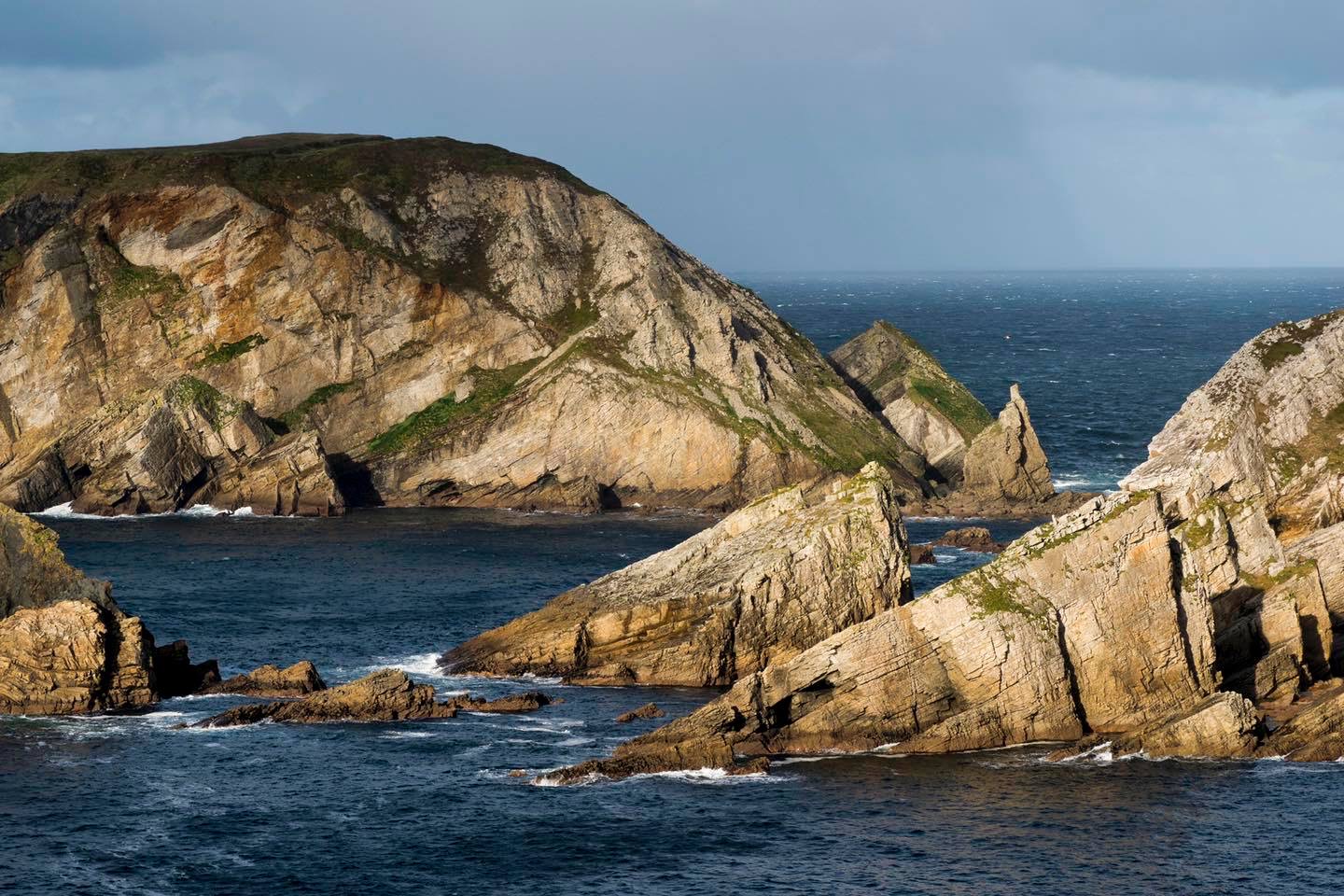 Ruige kliffen aan zee met steile, rotsachtige formaties rijzen op boven de diepblauwe oceaan en belichamen de aantrekkingskracht van een rondreis Ierland. De kliffen zijn versierd met stukken gras, geaccentueerd door zonlicht tegen een bewolkte lucht en verre regen. De contrasterende texturen creëren een dramatisch Iers kustlandschap. | Echt Ierland Vakanties