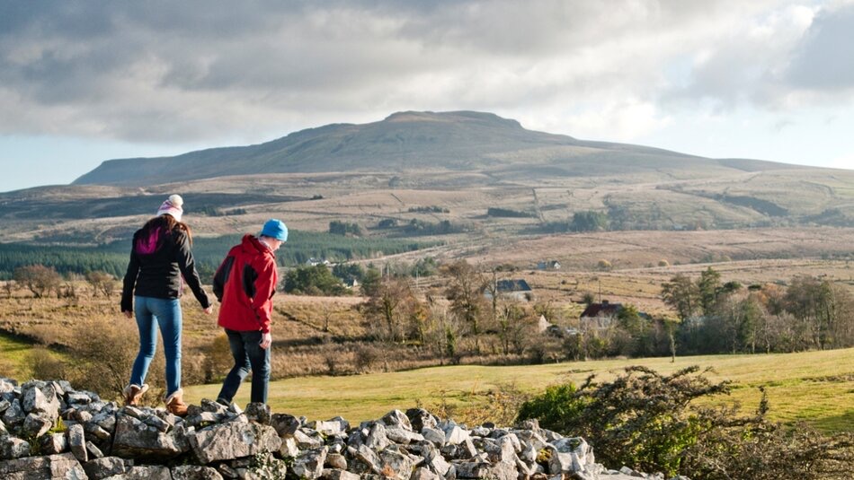 Twee mensen met jassen en hoeden lopen op een rotsachtig oppervlak op de voorgrond, wat de geest van Ierland vakanties belichaamt. Ze kijken uit over een uitgestrekt landschap van velden en verspreide gebouwen. Een grote heuvel domineert de achtergrond onder een bewolkte lucht, wat suggereert dat het een frisse, schilderachtige dag op het platteland is. | Echt Ierland Vakanties