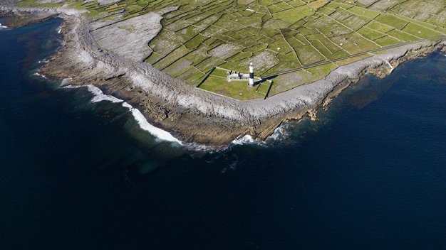 Luchtfoto van een ruige kustlijn tijdens een vakantie in Ierland, met een vuurtoren aan de rand, omringd door groene lappendekenvelden en stenen muren. De donkerblauwe zee contrasteert met de rotsachtige kust en creëert witte golven waar ze elkaar ontmoeten. Het tafereel geeft een sereen, geïsoleerd landschap weer. | Echt Ierland Vakanties