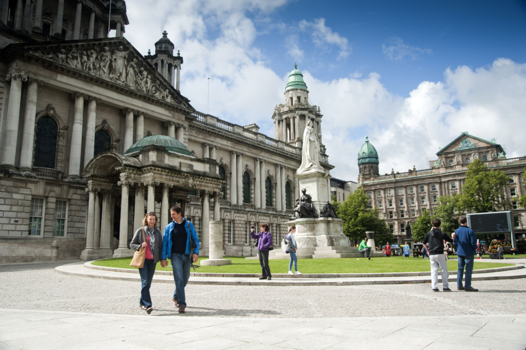 Mensen lopen in de buurt van het stadhuis van Belfast, een groot gebouw met een neoklassieke gevel en groene koepels, onder een gedeeltelijk bewolkte lucht. Een wit marmeren standbeeld staat op het gazon. Veel mensen die op vakantie zijn in Ierland verkennen het gebied terloops en benadrukken de aantrekkingskracht ervan als een historisch en architectonisch monument. | Echt Ierland Vakanties