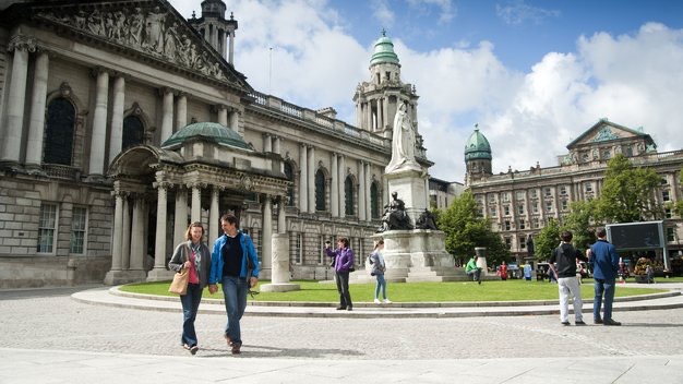 Een historisch stenen gebouw met zuilen en een groene koepel staat op de achtergrond, perfect voor degenen die genieten van rondreis Ierland. Een standbeeld staat prominent op een grasveld vooraan. Verschillende mensen dwalen rond en maken foto's, terwijl er een licht bewolkte lucht boven hangt. | Echt Ierland Vakanties