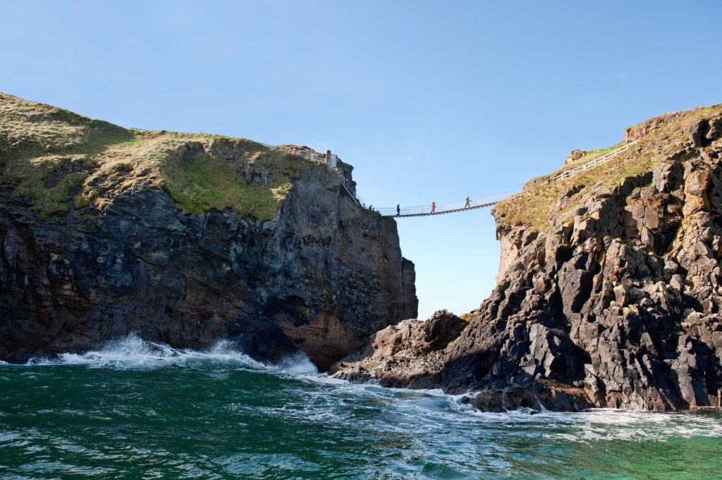 De afbeelding legt een prachtig tafereel vast van een rondreis Ierland, met een touwbrug die ruige kliffen over de oceaan verbindt. Meerdere mensen steken de brug over, bovenop met gras bedekte kliffen. Beneden is de zee diepgroen met golven, en boven is de lucht helder en blauw, wat een perfecte zonnige dag in Ierland belichaamt. | Echt Ierland Vakanties