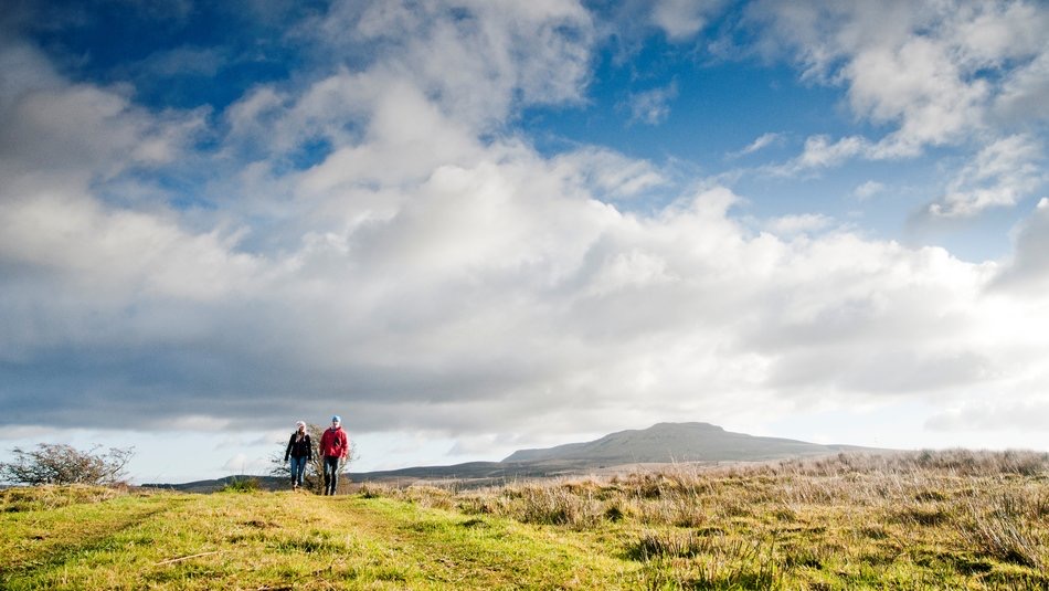 Twee mensen wandelen op een graspad onder een uitgestrekte, bewolkte lucht. De een draagt een rood jasje en de ander een blauw. In de verte, een eenzame berg gedeeltelijk verduisterd door wolken, hint naar de avonturen die je beleeft tijdens een betoverende vakantie in de open landschappen van Ierland. | Echt Ierland Vakanties
