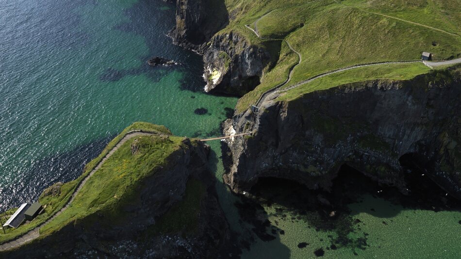 Luchtfoto van een smalle touwbrug die twee rotsachtige kliffen verbindt over helder turquoise water, een iconisch gezicht op een vakantie in Ierland. De met gras begroeide kliffen hebben wandelpaden en aan één kant is een klein, donker gebouw zichtbaar. De kalme zee eronder geeft een hint van de diepte met zijn donkere vlekken. | Echt Ierland Vakanties