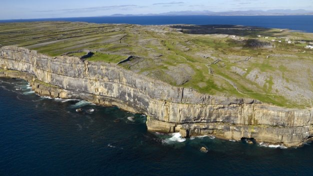 Luchtfoto van ruige, steile kliffen op Inis Mór in Ierland, begrensd door diepblauwe oceaan golven. Perfect voor een rondreis Ierland, de kliftoppen zijn grasachtig en oneffen, en strekken zich uit tot in de verte onder een licht bewolkte lucht. Spaarzaam groen contrasteert met het rotsachtige landschap. | Echt Ierland Vakanties
