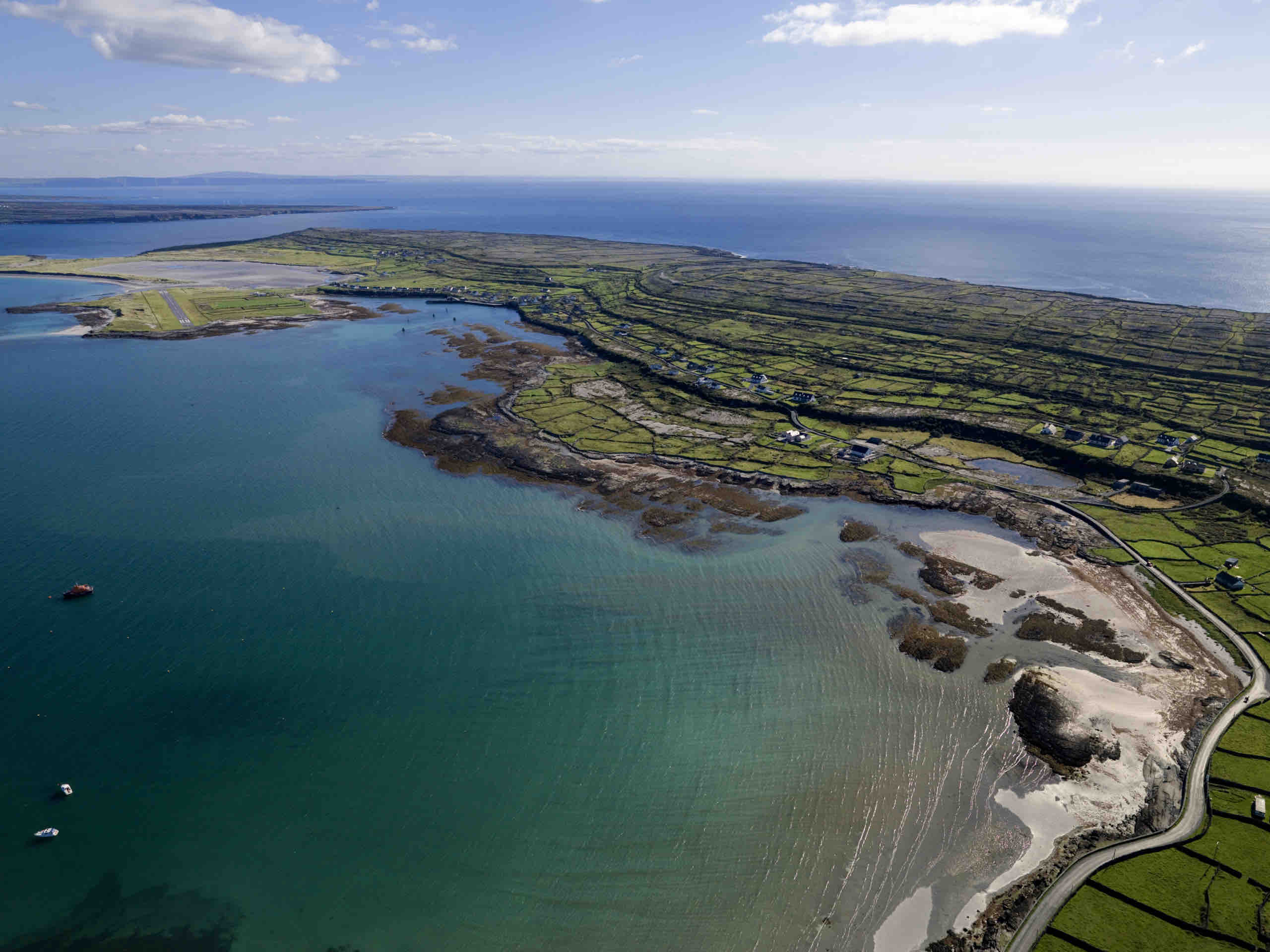 Luchtfoto van een eilandkustlijn in Ierland met helder turquoise water en een mix van zandige en rotsachtige oevers. Het landschap, dat doet denken aan een rondreis Ierland, bevat lappendekenvelden met stenen muren, verspreide gebouwen en een kronkelende weg die leidt naar de uitgestrekte, kalme zee onder een gedeeltelijk bewolkte lucht. | Echt Ierland Vakanties