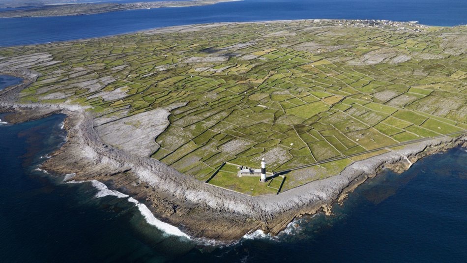 Luchtfoto van een kustlandschap in Ierland, perfect voor een rondreis Ierland, met groene velden die worden gescheiden door stenen muren. Een vuurtoren staat op de rotsachtige kustlijn met golven die er zachtjes tegenaan slaan. De oceaan strekt zich uit tot aan de horizon en ontmoet ver land onder een helderblauwe lucht. | Echt Ierland Vakanties