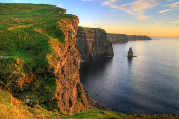 Steile, met gras begroeide kliffen rijzen boven kalme oceaanwateren uit bij zonsondergang tijdens een rondreis Ierland, met een eenzame verticale rotsformatie nabij de kust. De horizon gloeit met warm oranje licht, dat geleidelijk verzacht tot een helderblauwe lucht, waardoor een sereen en majestueus landschap ontstaat. | Echt Ierland Vakanties