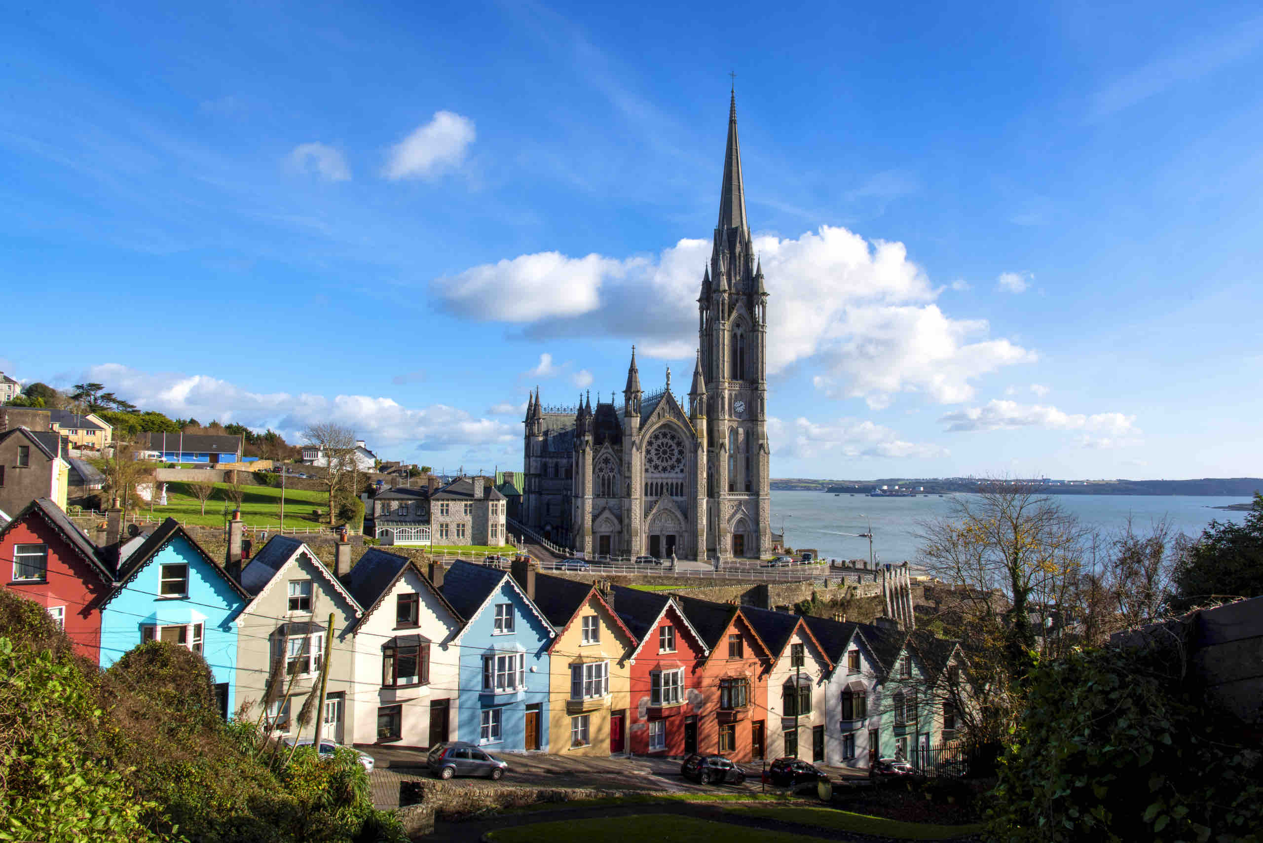 Kleurrijke huizen staan langs een straat op de voorgrond, die leidt naar een prachtige gotische kathedraal met een hoge torenspits, een must-see op elke vakantie in Ierland. De kathedraal kijkt uit over een schilderachtige haven, omlijst door groen en een helderblauwe lucht met donzige witte wolken. | Echt Ierland Vakanties